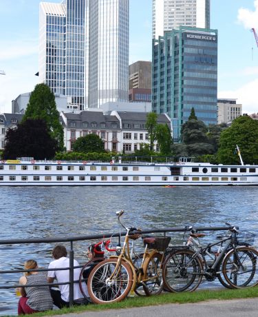 Fahrräder und am Ufer sitzende Menschen, dahinter die Skyline